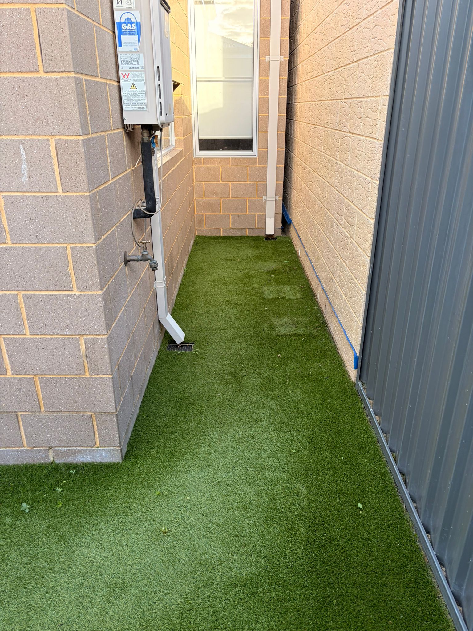 Narrow Atwell side-return passage before cleaning, the synthetic turf strip flat and patchy along the walked centre line with leaf debris at the edges